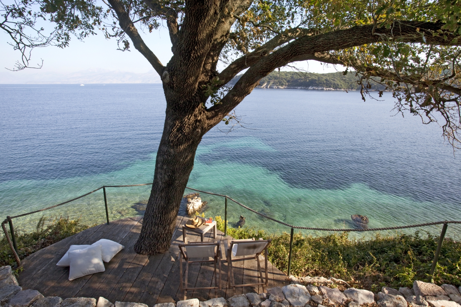 Beach side balcony with deck chairs, coffee table, cushions, tree and sea view at the Odysseus Estate on Corfu, Greece