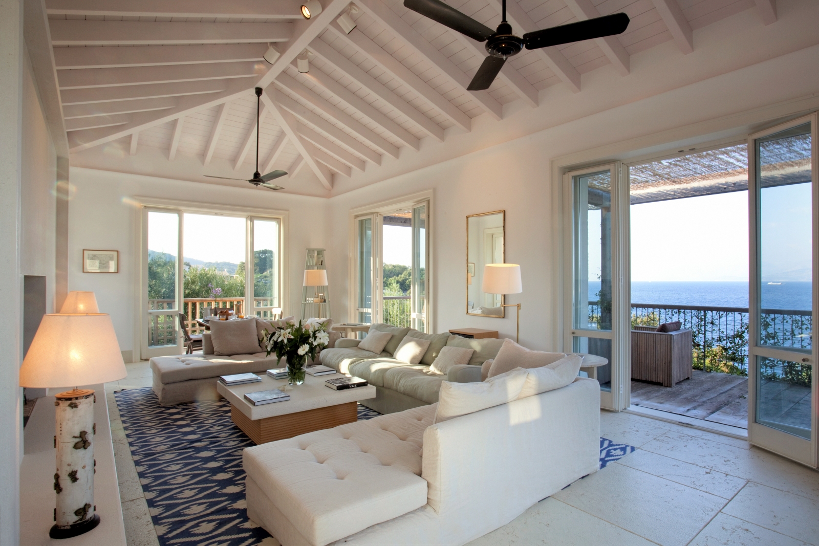 Living room with sofas, coffee table, flowers, lamps, fans, French doors and sea view at the Odysseus Estate on Corfu, Greece