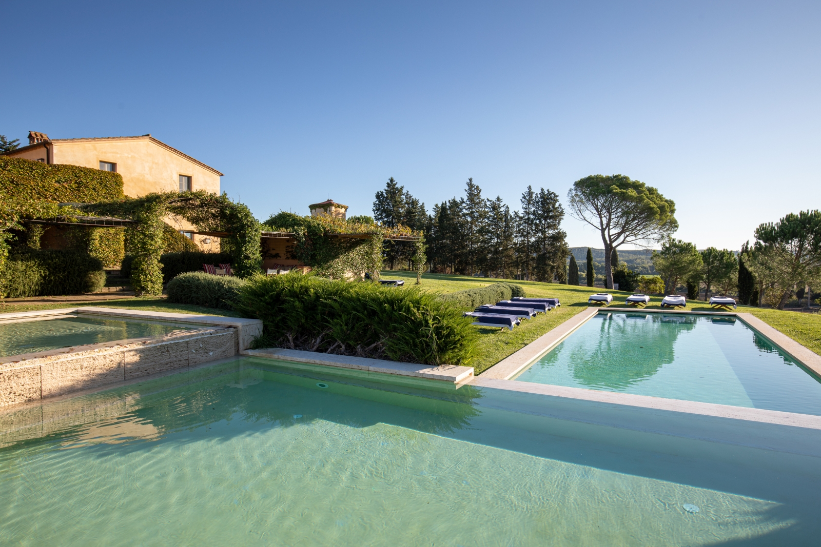 Large swimming pool in garden with sun loungers, grass, hedges & trees at Il Serratone in Tuscany, Italy