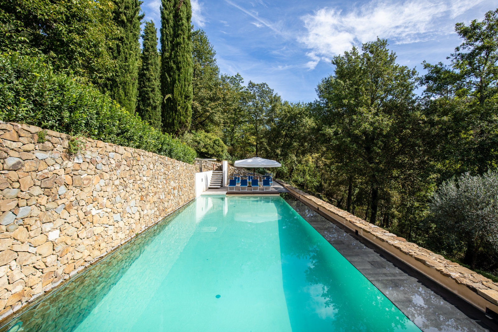 Pool with sun loungers, umbrella, stone wall and trees at Villa Baciata in Tuscany, Italy