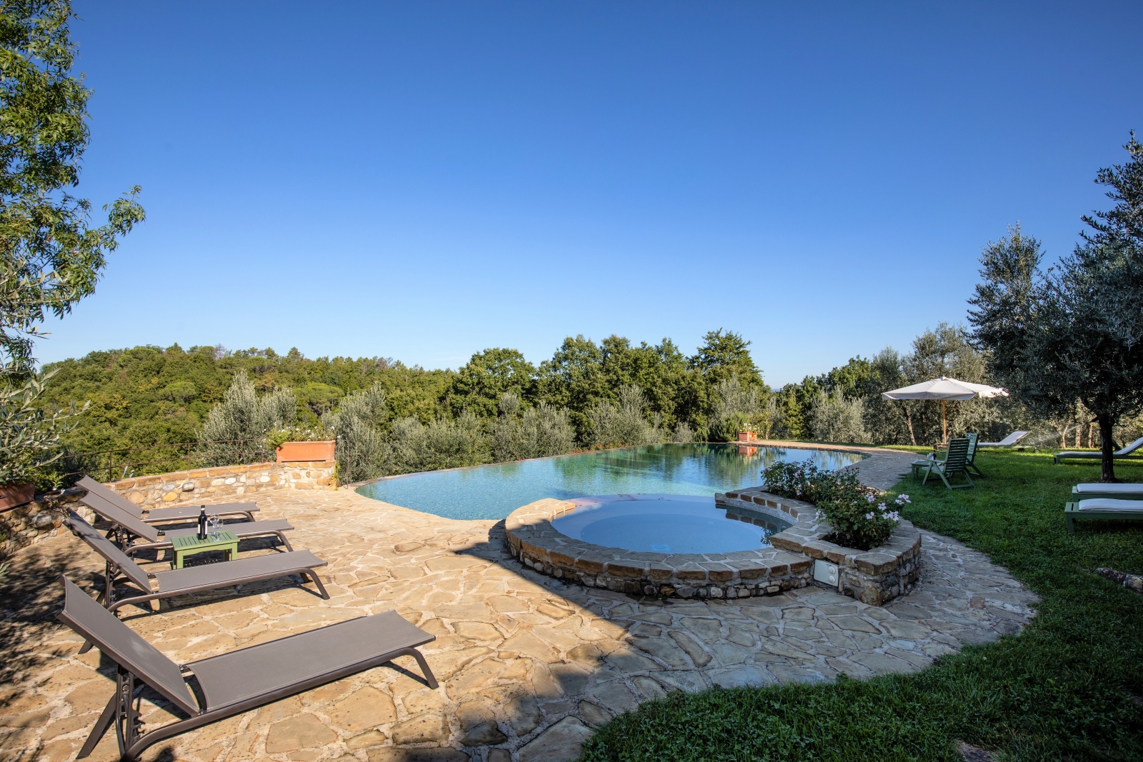 Patio next to pool with sun loungers, table, wine, umbrella and countryside view at Villa Giuliana in Tuscany, Italy