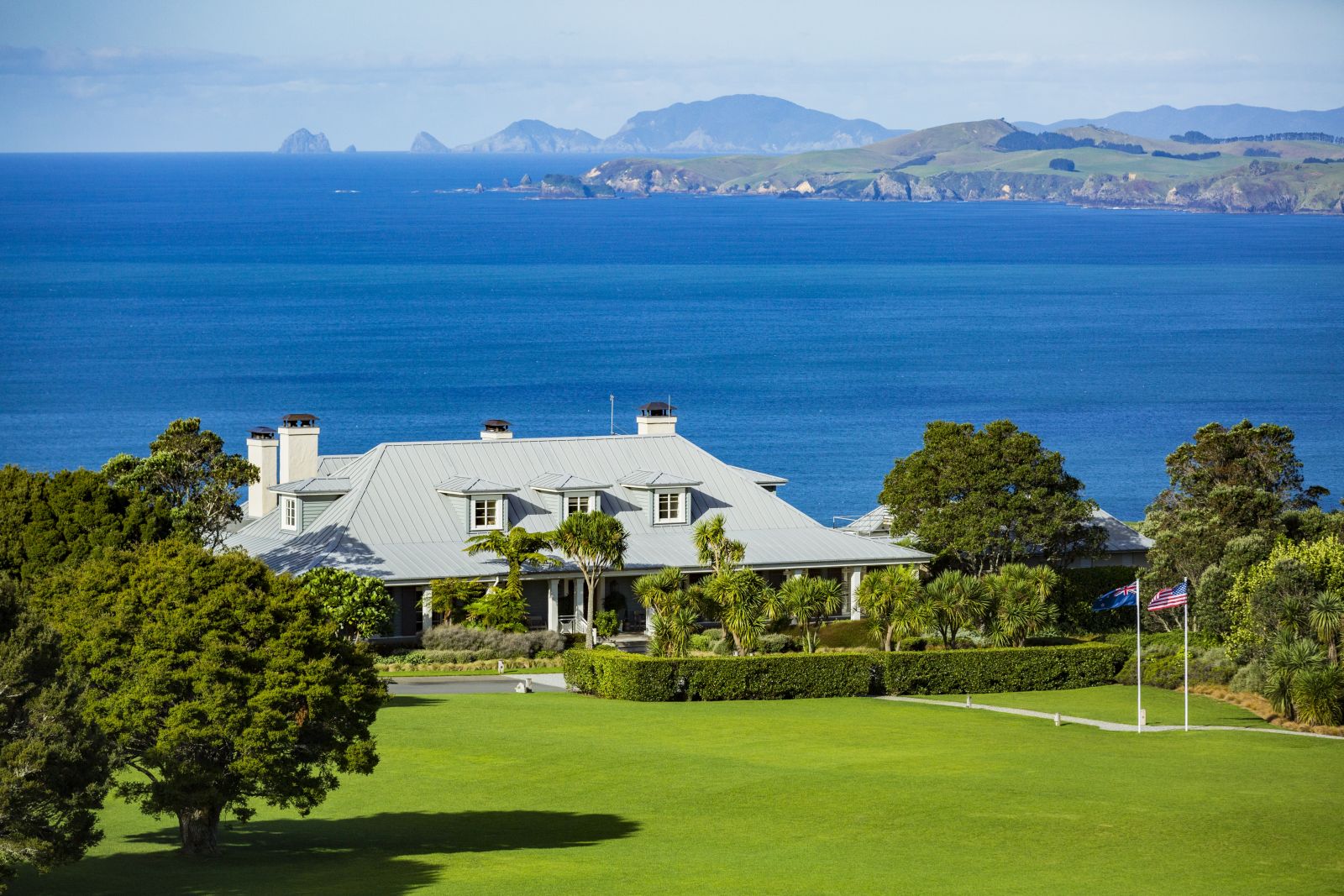 Exterior shot of Kauri Cliffs Lodge in New Zealand with green lawns in front and the Pacific Ocean and mountains in the background