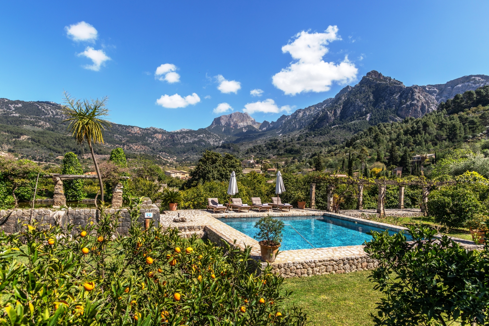View of pool with sun loungers, umbrellas, stone pillar arbour, mountains and orange tree at Can Canals in Mallorca, Spain