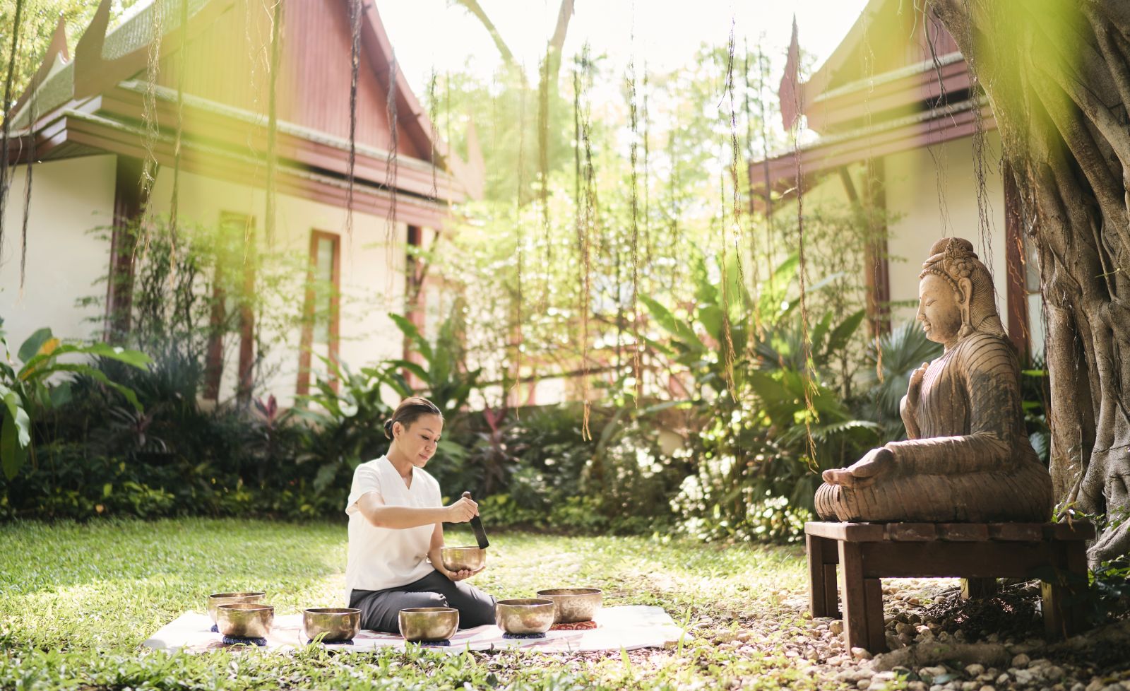 Lady performing Tibetan Sound healing bowls in a garden in front of a buddha statue at Luxury resort Chiva Som